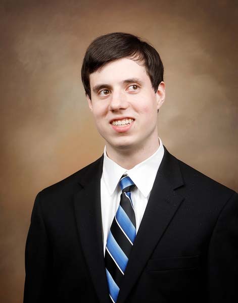 Portrait of Timothy Jones. Timothy can be seen from mid torso up and is sitting in front of a brown background. He is wearing a black suit, white collared shirt, and a blue and black diagonally striped tie.